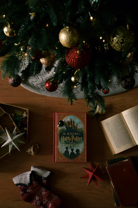 Books and a box of Christmas decorations on a wooden floor below a Christmas tree. ‘Harry Potter and the Philosopher’s Stone’ is the book in the centre of the image. 