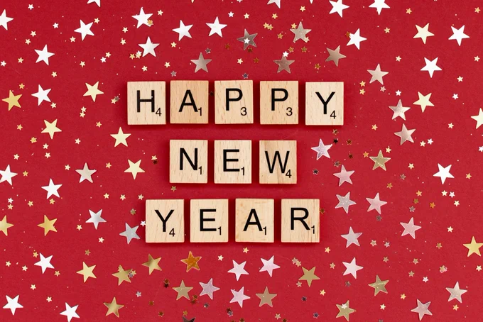 Scrabble tiles spelling out ‘Happy New Year’ on a red background decorated with gold and silver stars.