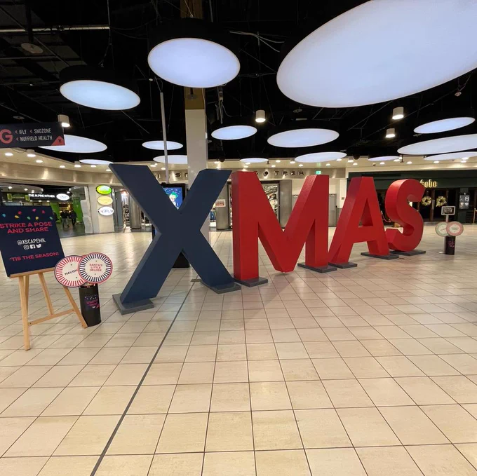 Large "XMAS" letters displayed in a spacious indoor area at Xscape Milton Keynes with circular white lights on the ceiling.