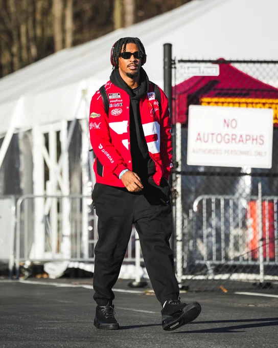 Photo of Jayden Daniels arriving at the stadium. He is wearing sunglasses, a red racing jacket and black pants.