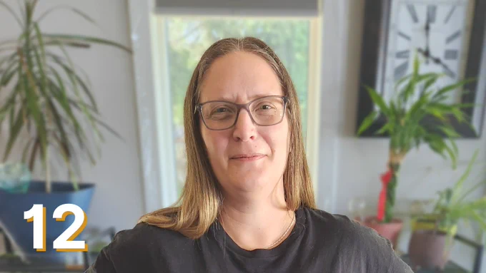 Patient partner Christina LaBerge, smiling. She seems to be standing in her home. A clock, a window and many plants can be seen behind her.