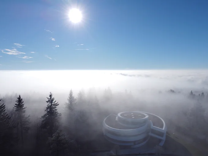 Das Haus der Astronomie im Wald eingehüllt in eine dünne Nebelschicht, darüber Sonne mit blauem Himmel