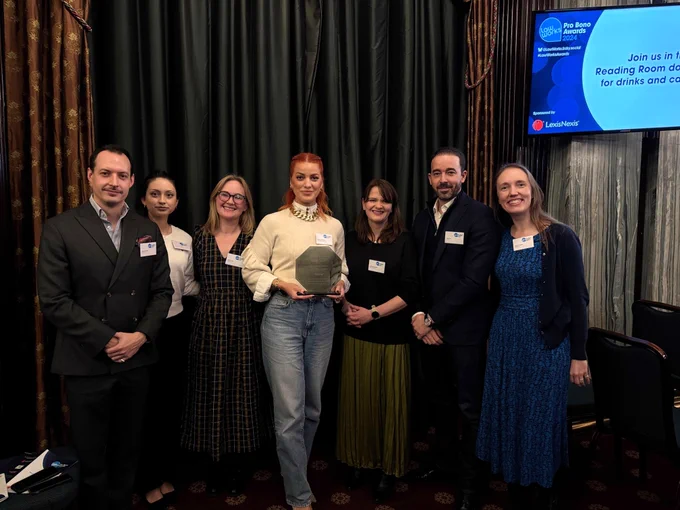 A group of seven people pose for a photo after accepting the Best International Pro Bono award on behalf of Reed Smith LLP, Amazon, Bank of America and Face Equality International. Matilda from FEI stands in the middle holding the glass award. Behind the group are elaborate draped curtains and a TV screen with the name of the award ceremony.