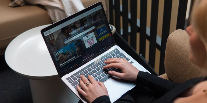 Person using a laptop displaying the UCAS website while sitting on a couch in a casual office lounge.