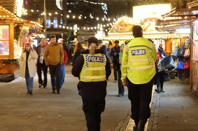 Two police officers can be seen wearing yellow high vis and police baseball caps walking through the Christmas markets in Sheffield. Stalls can be seen the left and right, lit up with Christmas lights.