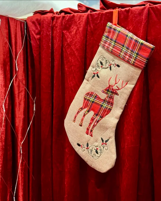 A Christmas stocking featuring a reindeer design hangs in front of a curtain of deep red velvet.
