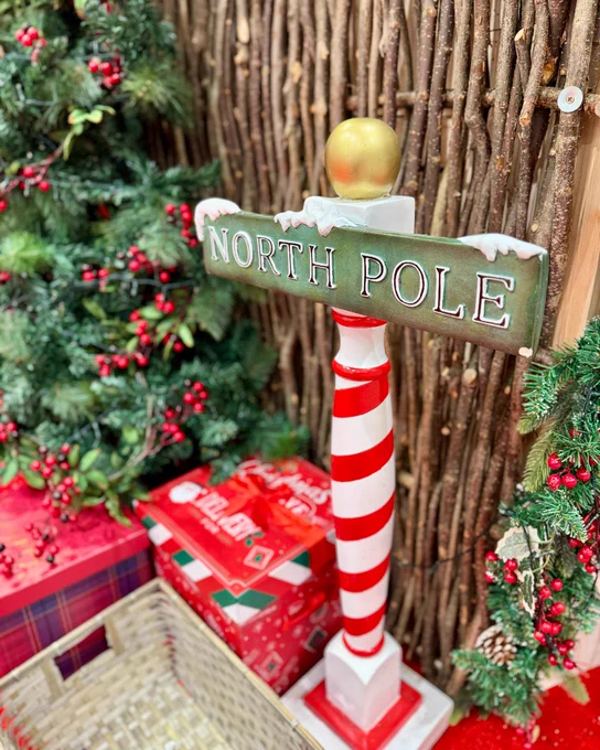 A festive display featuring a "North Pole" signpost with a red and white striped pole, surrounded by Christmas trees and gifts.