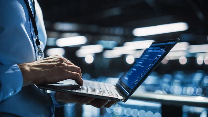A professional using a laptop with data charts on the screen in a dimly lit industrial environment.