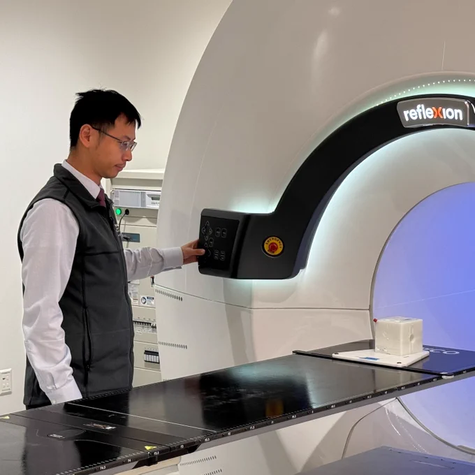 Dr. Cai in a white long sleeve shirt and black vest standing at a radiation treatment machine.