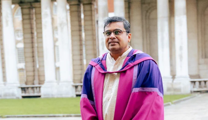 Sachan Das, an Indian man wearing glasses and a pink robe with purple detailing, is standing outside a grand building.