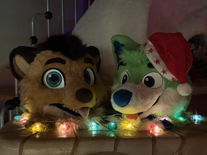 Two fursuit heads, a meerkat head on the left and a green husky head with a Christmas hat on the right with lights decorating the scene