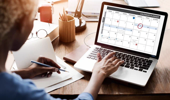 Stock image of a woman sitting at a laptop, one hand is writing in a notebook while another types, with a calendar onscreen.