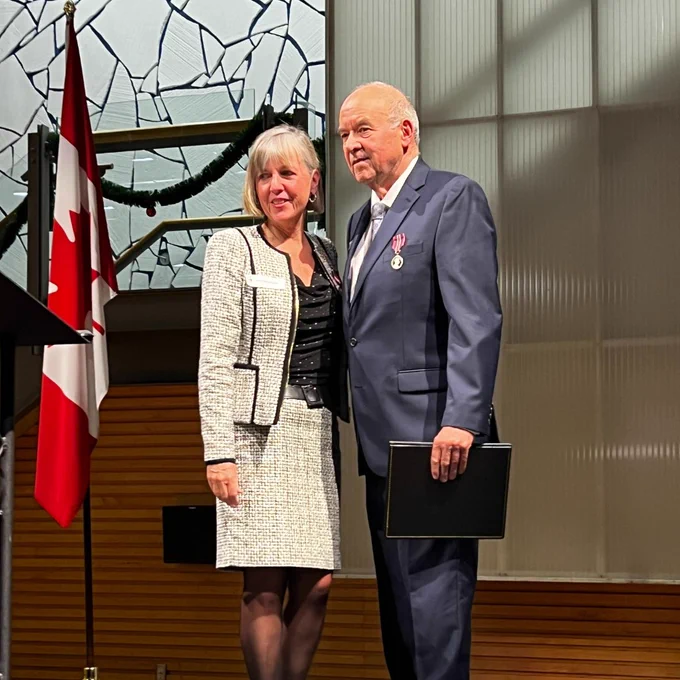 A photo of two people posing for a photo on a stage. There is a Canadian flag nearby.