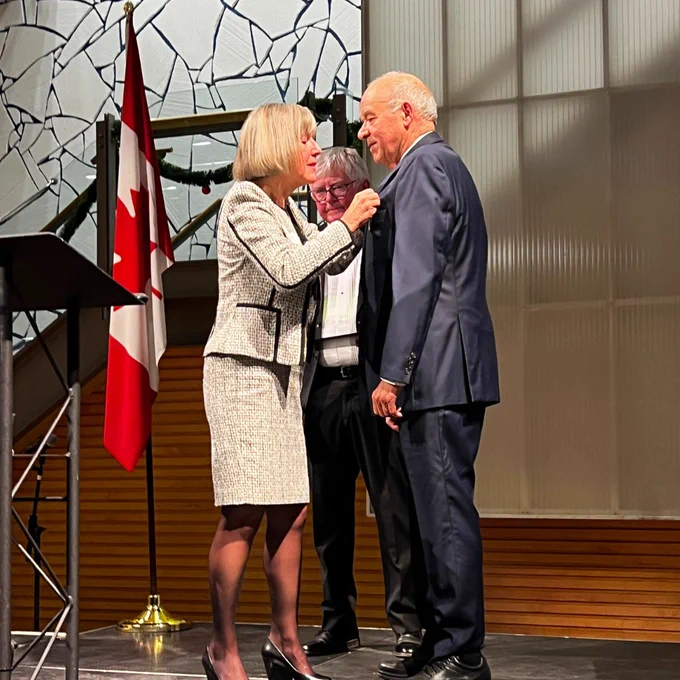 A photo of a woman posting a pin to a man's jacket. They are standing on a stage, which also features a podium and Canadian flag.