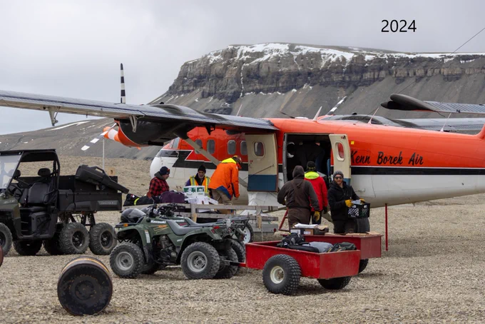Des personnes déchargent des fournitures d’un avion et les placent sur des véhicules tout-terrain munis de remorques. Un escarpement recouvert de neige est visible à l’arrière-plan.