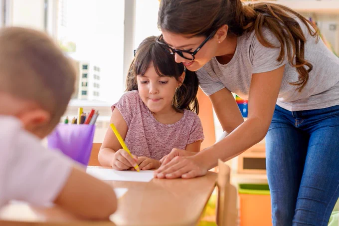 Teacher assisting a child with writing in a classroom, another child present.
