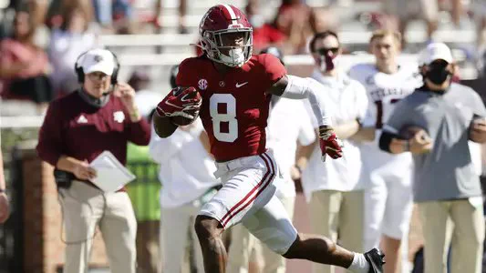 John Metchie III carries the football as the opposing team on the sideline watches him run by them during an Alabama Crimson Tide football game.