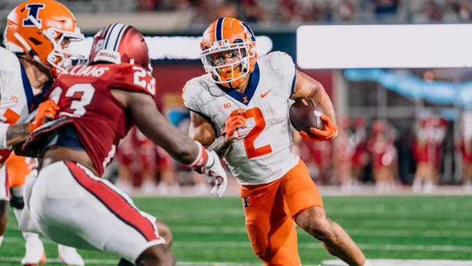 Chase Brown carries the football as a teammate blocks for him and a defender tries to get off the block during an Illinois Fighting Illini football game.