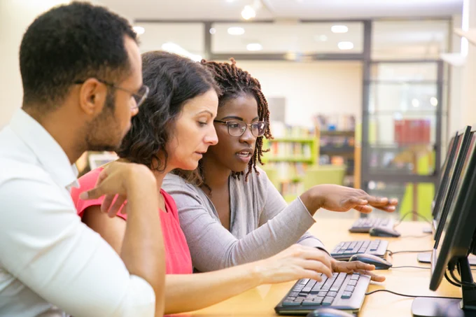 Stock image of three individuals seated next to one another at a row of computers, talking while gesturing at the middle computer as the seated woman types.