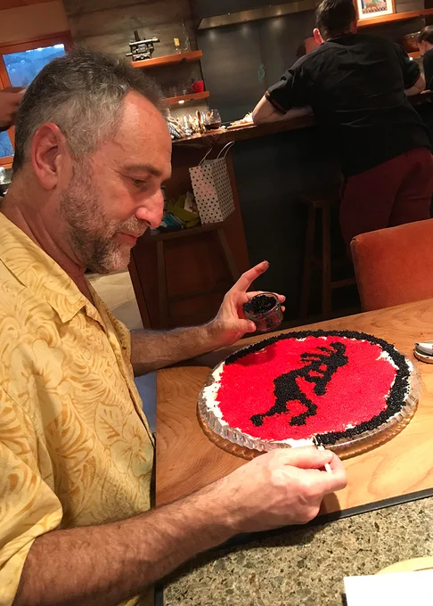Working at a kitchen table, a man in a short-sleeved dress shirt creates a design of Kokopelli, a silhoutted figure with a feathered headdress playing flute, out of red and black caviar on a circular plate. 