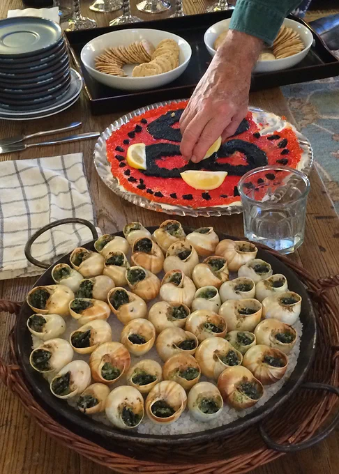 A hand holding a cracker reaches into a plate of red and black caviar, garnished with lemon slices. In front of the caviar is a metal paella pan filled with white rock salt and pale brown snail shells, each filled with a dark green mixture.