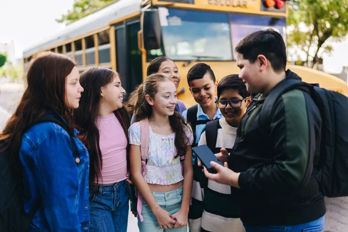 Middle school students gather together in front of a school bus outside a school.