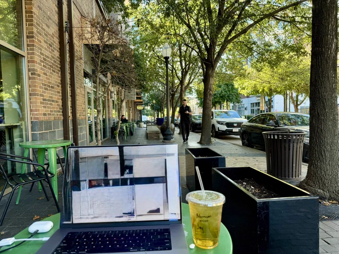 Picture from sitting at a sidewalk cafe with an iced tea and a laptop on the table in front of you, a guy walking up the sidewalk, parked cars at the curb, and mid-rise density all around.