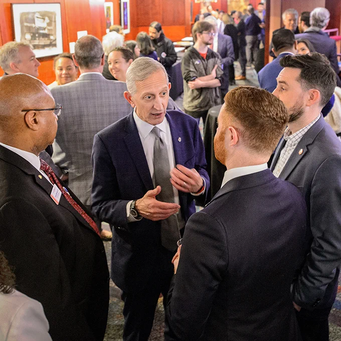 Mayor Wilson stands in the middle of a group of people, talking to them, while at a large reception.