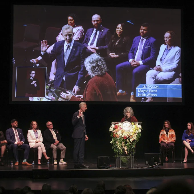 Mayor Wilson stands at the center of a theater stage with his right hand raised and facing Judge Ortega. A line of elected officials are also on the stage.