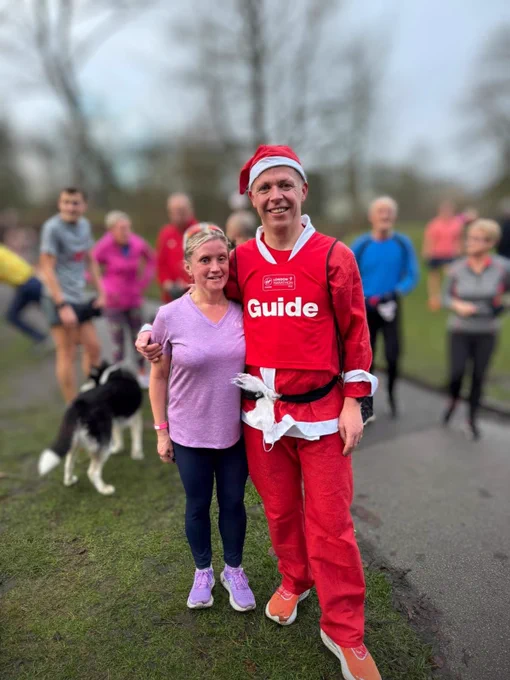 The image shows a man and a woman standing together outdoors on a grassy area. The man is wearing a red Santa hat and a red outfit with white trim, resembling a Santa Claus costume. His outfit has a label on the chest that reads "Guide" in large white letters, and above it, there is a logo with the text "London Marathon." The woman next to him is wearing a light purple short-sleeved shirt and dark leggings. She has her arm around the man's waist, and they are both smiling at the camera. In the background, there are several other people, some wearing athletic clothing, and a black and white dog is visible. The setting appears to be a park or an open outdoor area with trees in the distance.