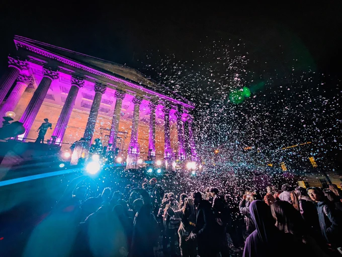 UCL portico lit up in purple. Students are gathered in front taking photos and looking up at the fake snow being sprayed. 