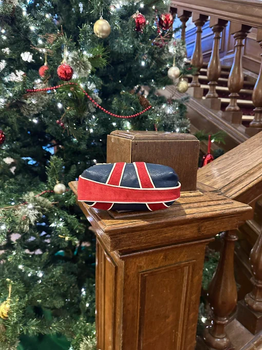 Capsule shaped clutch bag sitting on a wooden bannister in front of a large Christmas tree. The bag is made from blue leather with blue and white details, inspired by the Union Jack flag.
