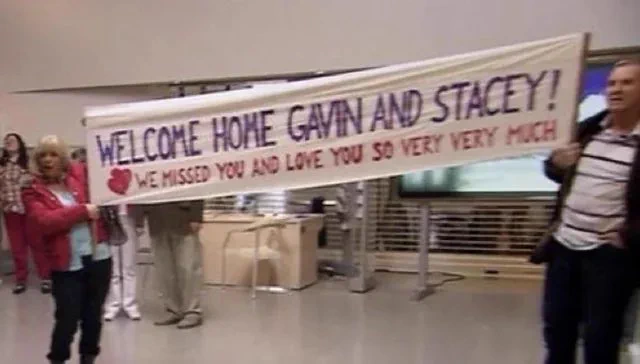 Pam and Mick holding a big banner saying, “Welcome home, Gavin and Stacey! We missed you and love you so very, very much.”