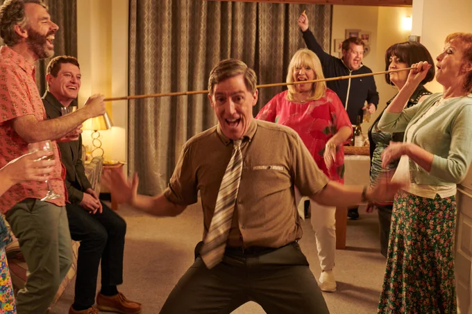 A lively party scene with the cast of Gavin and Stacey smiling and dancing in a warmly lit living room. Bryn, in the centre, performs a limbo under a stick held by Gwen and Jason.