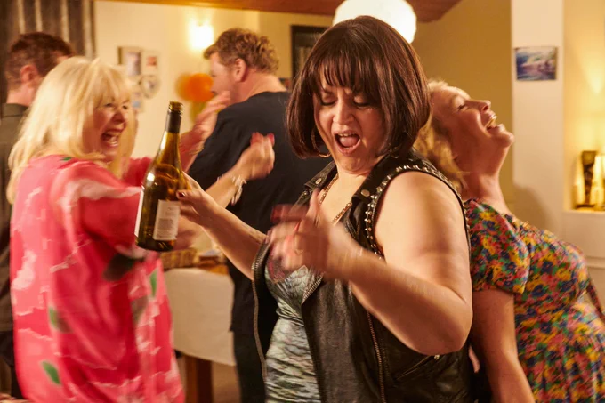 Pam, Nessa and Stacey enjoying a lively party with dancing and laughter, Nessa holding a bottle of wine.