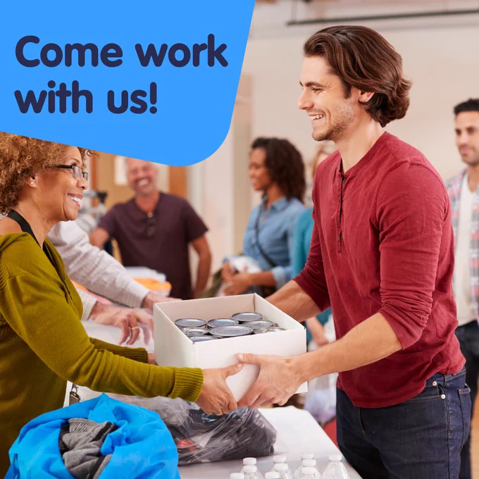 Come work with us! Two volunteers smiling and interacting at a community center, with others working in the background.