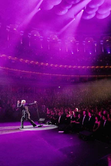 Robbie Williams singing on stage at a live concert, with an audience watching attentively in a venue with ornate, tiered balconies illuminated by purple stage lights.