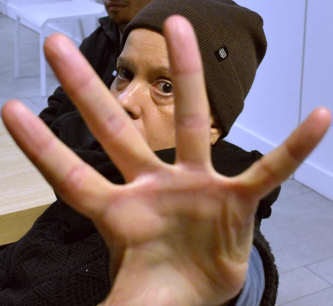 Primer plano de la palma de una mano, con los dedos extendidos. A través de ellos vemos los ojos de una mujer que lleva un gorro marrón y viste un jersey negro.