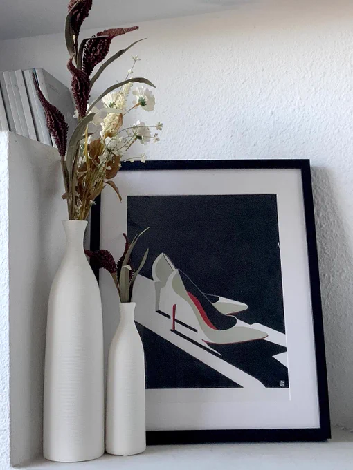 Dibujo enmarcado de unos zapatos de tacón de color blanco, junto a unos jarrones de color blanco con flores blancas. Pared de fondo y estantería en yeso blanco.