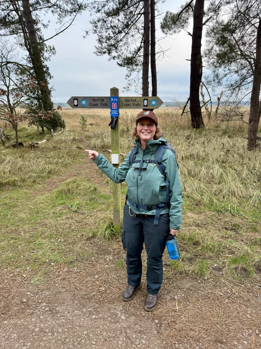 A woman (me) wearing hiking gear pointing in front of a Norfolk Coast Path fingerpost