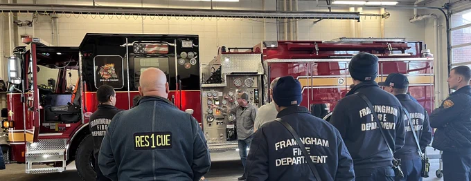 Image of 6 firefighters watching an instructor pointing to the side panel on a firetruck.  