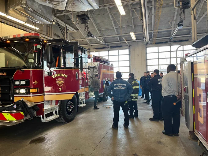 Group of firefighters watching an instructor as he stands in front of a red firetruck with its driver side door open as he points out the truck's features.