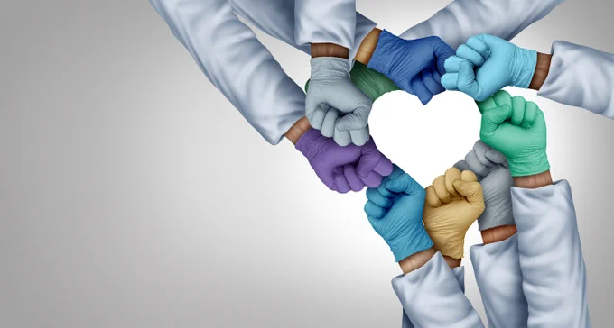 A group of healthcare workers wearing medical scrubs and colored gloves form a heart shape with their fists against a gray background.