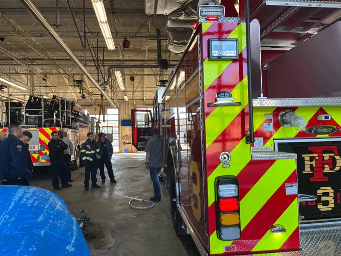 Group of 7 firefighters watching an instructor as he stands in front of a red firetruck with its driver side door open as he points out the truck's features.