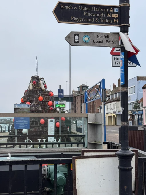 A fingerpost in Wells harbour pointing to the Norfolk Coast Path, outer harbour, pinewoods, beach and playground. Behind it is a Christmas Tree made out of crab pots which was originally conceived in lockdown.