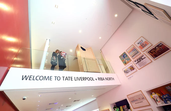 Two visitors standing on the mezzanine at Tate Liverpool   RIBA North, which overlooks the shop. There is a sign just below the visitors which says 'WELCOME TO TATE LIVERPOOL   RIBA NORTH'. There are framed prints hanging on the wall on the right hand side.