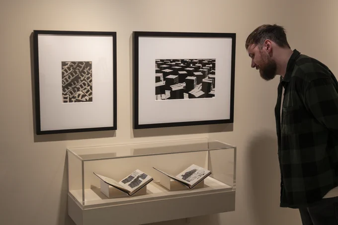 A visitor is standing and observing artworks displayed in a glass case on the wall showcasing open sketchbooks. There are two framed artworks on the wall above the case. 