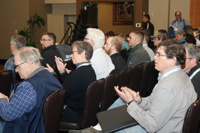 Audience members seated in rows at a conference, clapping and looking towards a speaker off-camera.