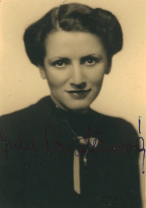 Vintage black and white portrait of a woman with a confident smile, wearing a dark outfit and styled hair. The photo is signed at the bottom.