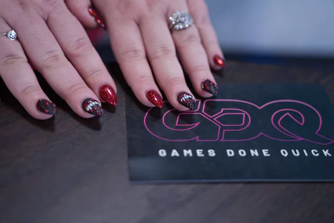 Image of nails painted in black and red, middle finger on both hands have vampire teeth on them. The hands are next to a GDQ business card